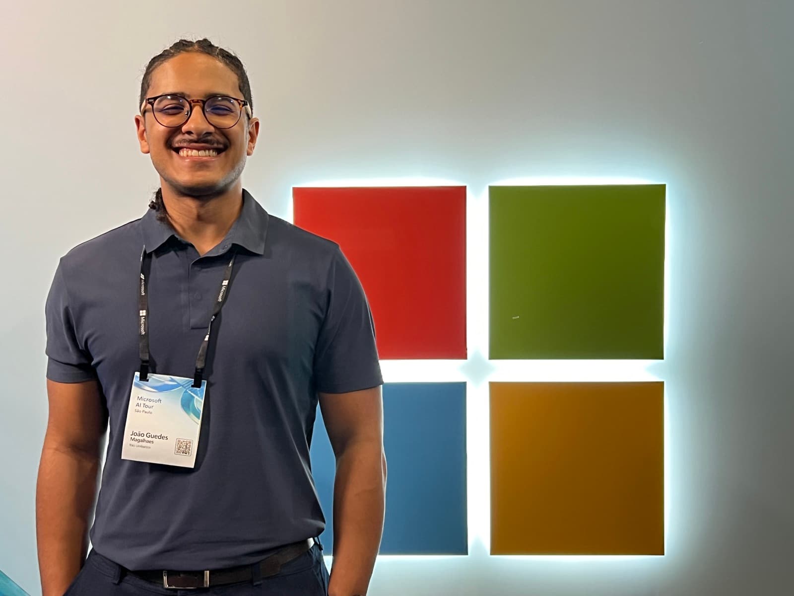 João Guedes sorrindo em frente ao logo da Microsoft, no evento Microsoft AI Tour São Paulo